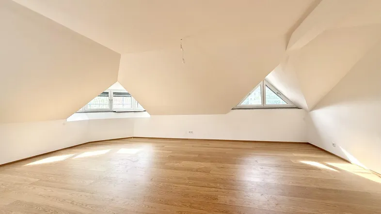 Heller Raum im Dachgeschoss mit schrägen Wänden, Holzboden und dreieckigen Fenstern.