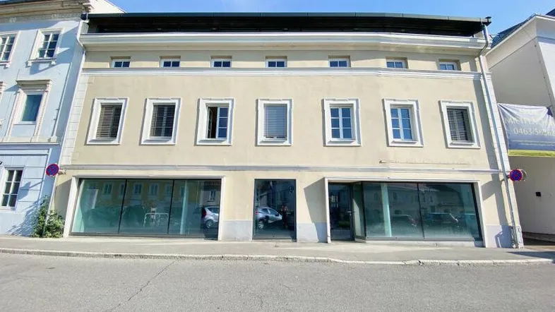 Fassade eines Geschäftsgebäudes mit großen Schaufenstern und oberen Wohnungen unter blauem Himmel.