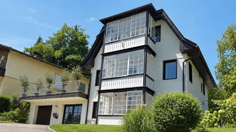 Weißes Haus mit dunklem Dach und mehreren verglasten Balkonen in grüner Umgebung.