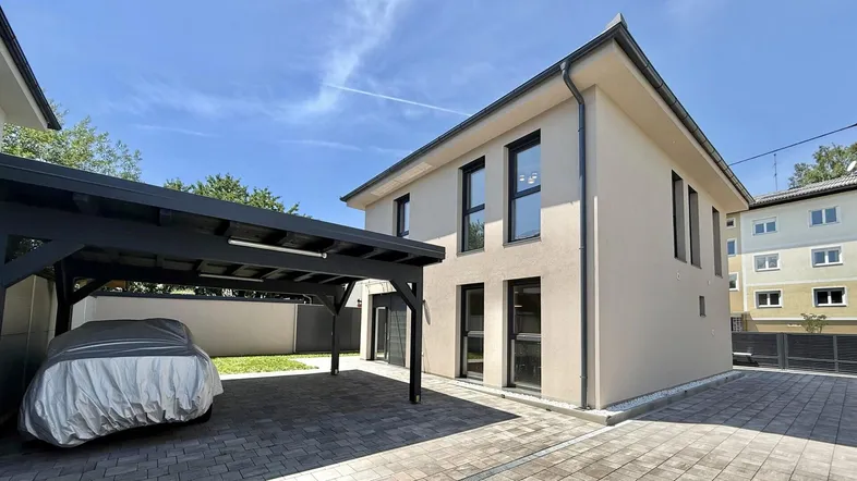 Modernes Haus mit beigefarbener Fassade und einem Carport für zwei Fahrzeuge auf gepflastertem Vorplatz.