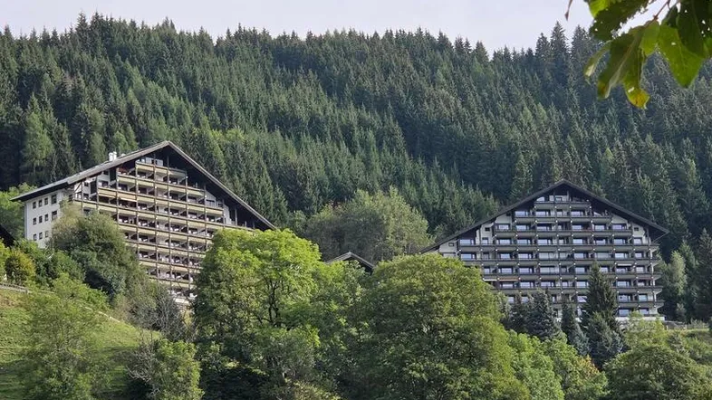 Große Apartmentgebäude in alpiner Landschaft, umgeben von dichtem Wald und grünen Hügeln.