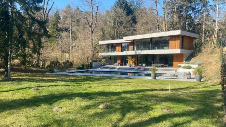 Modernes Haus mit großem Pool und Terrasse, umgeben von viel Grün und Bäumen.