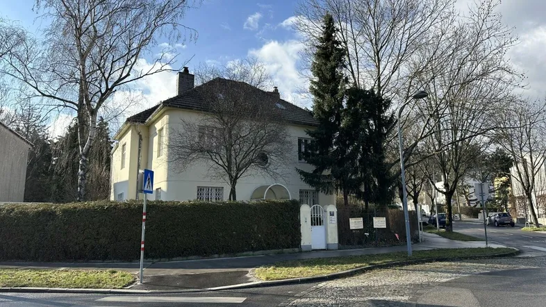 Gelbe Villa mit Hecke und Bäumen an einer Straßenecke unter blauem Himmel.