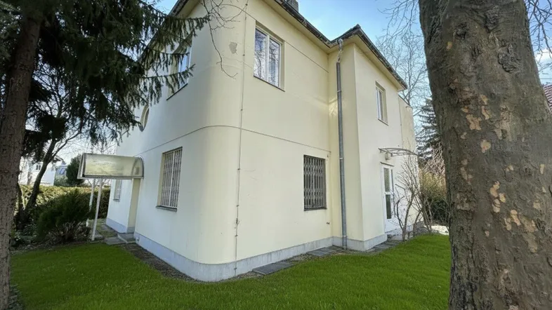 Seitenansicht des Gebäudes mit heller Fassade, umgeben von Rasenflächen und Bäumen.