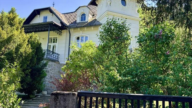 Historische Villa mit markantem Turm und schmiedeeisernem Zaun, umgeben von üppigem Grün.