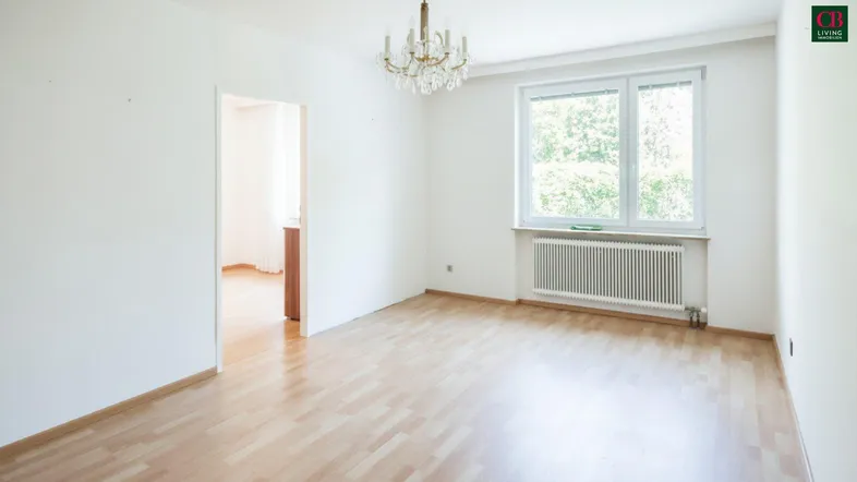 Helles, leeres Zimmer mit Parkettboden, großem Fenster und Kronleuchter, bietet Blick ins Grüne.