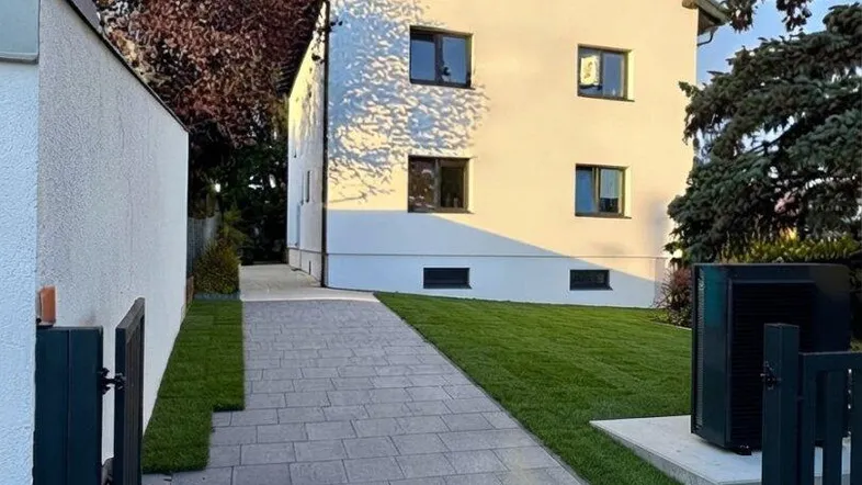 Außenansicht des modernen Gebäudes mit gepflastertem Weg, grünem Rasen und Bäumen unter strahlendem Himmel.