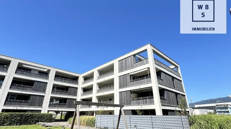 Modernes Mehrfamilienhaus mit klarer Architektur und einem kleinen Spielplatz im Vordergrund unter blauem Himmel.
