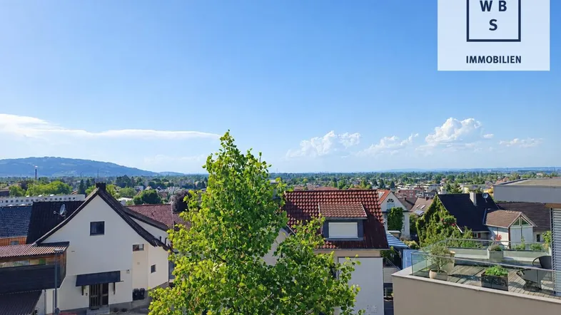 Weitblick über eine Wohnsiedlung mit grünen Bäumen und Bergen am Horizont unter blauem Himmel.
