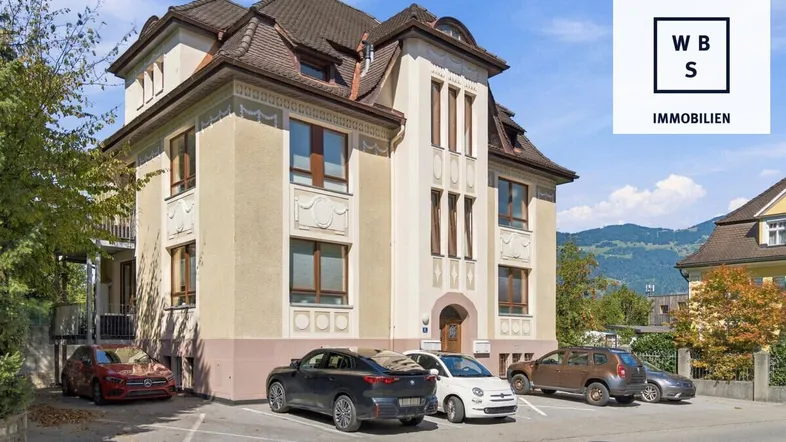 Historisches Gebäude mit klassischer Architektur, Parkplätzen und Bergblick im Hintergrund.