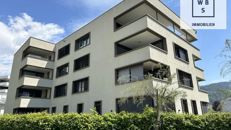 Modernes Mehrfamilienhaus mit klaren Linien und mehreren Balkonen, umgeben von grüner Hecke.