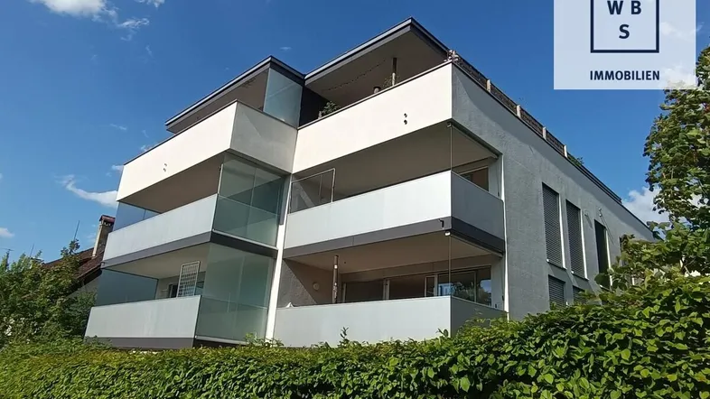 Modernes Mehrfamilienhaus mit Balkonen und gepflegter Hecke unter blauem Himmel.