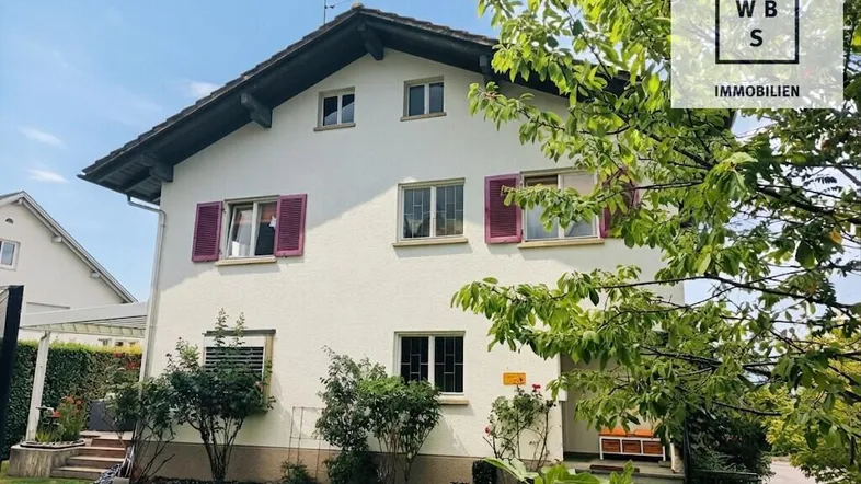 Helles Einfamilienhaus mit weißer Fassade, roten Fensterläden und gepflegtem Vorgarten.