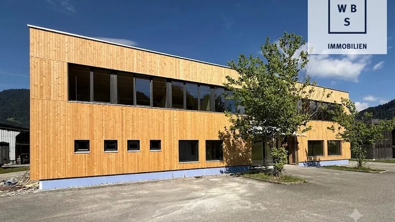 Modernes Gebäude mit Holzfassade und großen Fenstern unter blauem Himmel, umgeben von Natur.