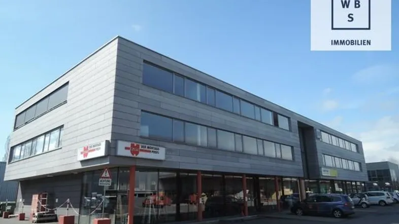 Modernes Geschäftsgebäude mit grauer Fassade, großen Fenstern und einem Parkplatz davor unter blauem Himmel.