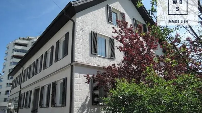 Mehrstöckiges Gebäude mit weißer Fassade, dunklen Fensterläden und einem Satteldach, umgeben von Bäumen.