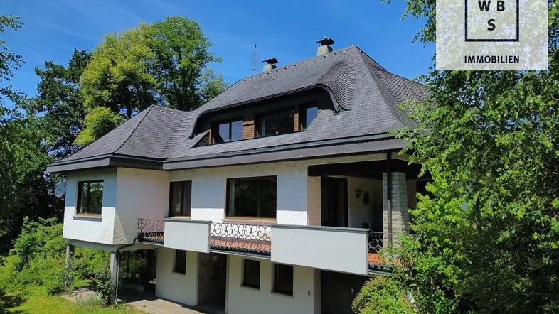 Freistehendes Haus mit markantem Dach und Balkon, umgeben von viel Grün und Bäumen.