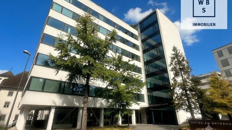 Modernes Bürogebäude mit Glasfassade und weißer Struktur, umgeben von Bäumen unter blauem Himmel.
