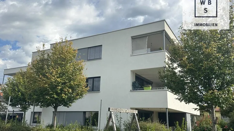 Moderne Wohnanlage mit weißer Fassade, Balkonen und grüner Bepflanzung unter bewölktem Himmel.