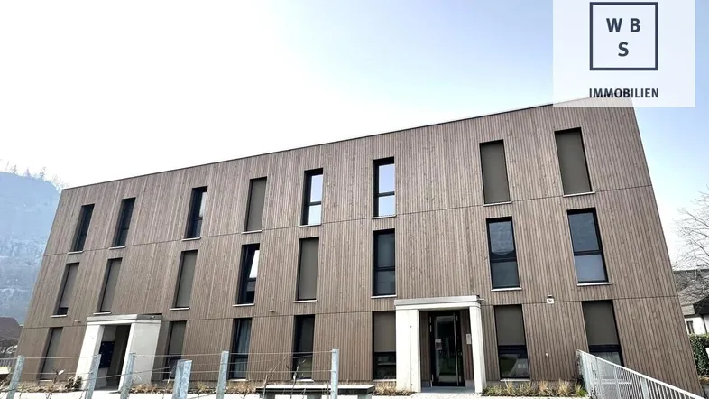 Modernes Mehrfamilienhaus mit Holzfassade und großen Fenstern in einer grünen Umgebung.