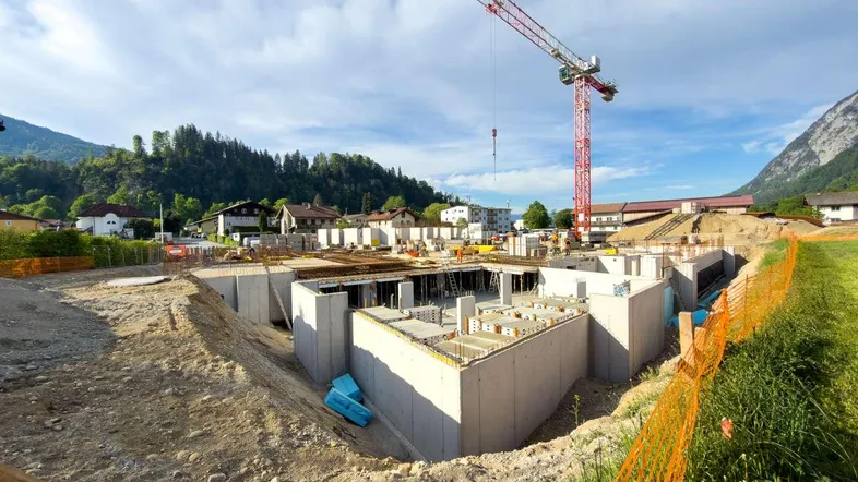 Baustelle mit bereits errichteten Betonfundamenten und einem Baukran vor bergiger Kulisse.
