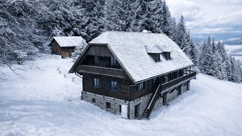 Traditionelles Holzhaus mit Steinfundament und schneebedecktem Dach in winterlicher Berglandschaft.