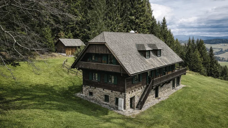 Traditionelles Holzhaus mit Steinsockel und Schindeldach, umgeben von Wald und Wiesen.