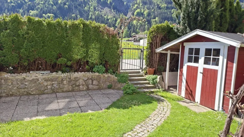 Gepflegter Gartenbereich mit Rasenfläche, Steinweg und einem roten Gartenschuppen vor bergiger Kulisse.
