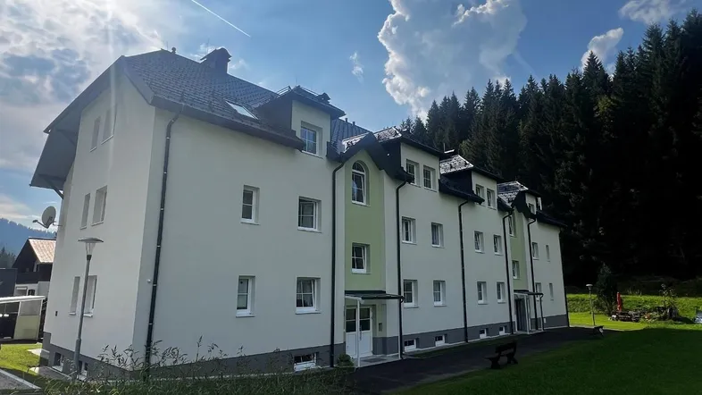 Mehrfamilienhaus mit heller Fassade und Satteldach, umgeben von Grünflächen und Wald.