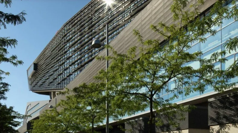 Modernes Bürogebäude mit Glasfassade und Bäumen im Vordergrund unter blauem Himmel.