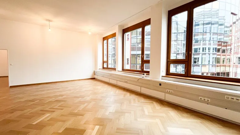 Heller, leerer Raum mit Fischgrätparkett und großen Fenstern, die viel Tageslicht hereinlassen.