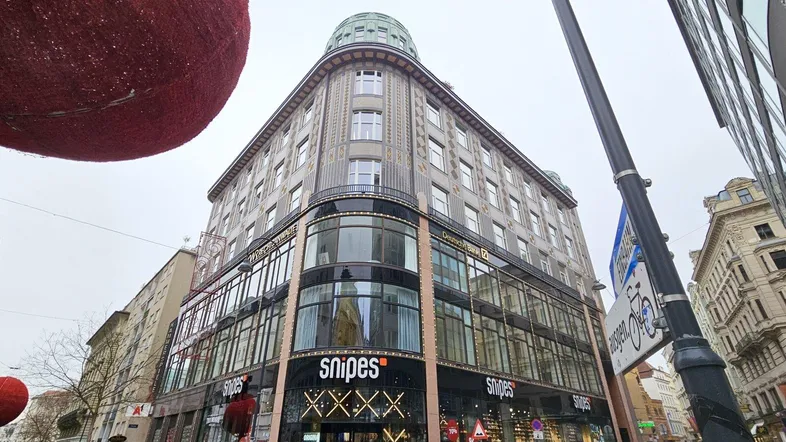 Großes Eckgebäude mit vielen Fenstern und Geschäften im Erdgeschoss, umgeben von städtischer Architektur.