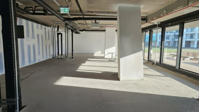 Geräumiger Büroraum im Rohbauzustand mit hohen Decken, sichtbaren Installationen und großen Fensterfronten.