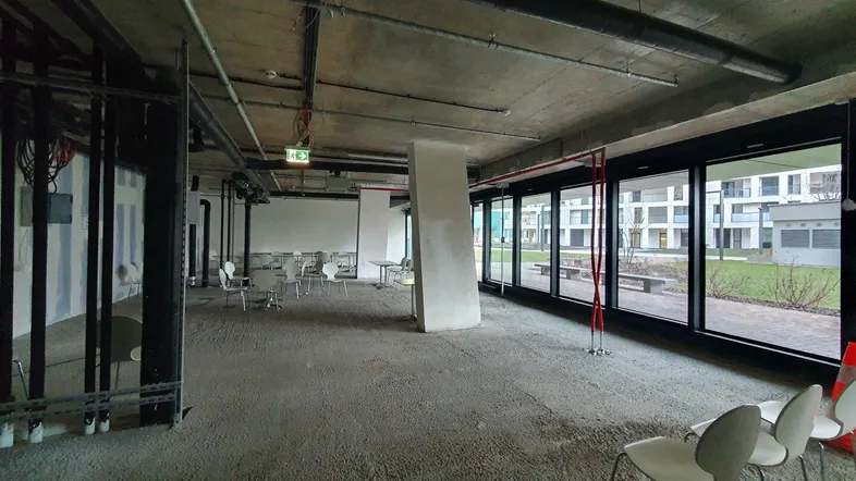 Großer, offener Innenraum im Rohbau mit Betonboden, sichtbaren Leitungen und großen Fenstern zum Außenbereich.