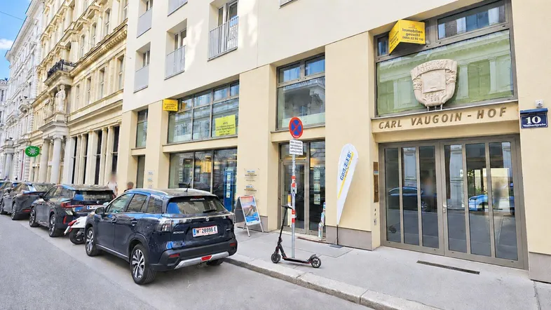 Fassade eines modernen Gebäudes mit Geschäften im Erdgeschoss und parkenden Autos auf der Straße.