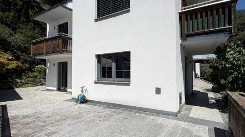 Moderne weiße Fassade eines Hauses mit großzügiger Steinterrasse und Holzbalkonen im Grünen.