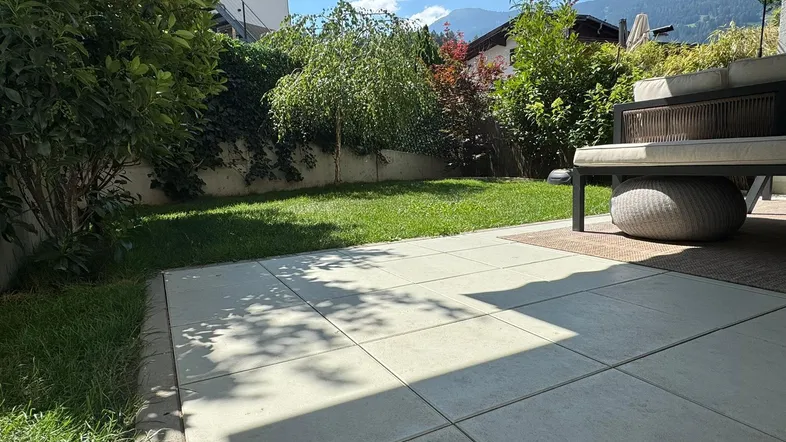 Gepflegter Gartenbereich mit Rasenfläche und einer sonnigen Terrasse, umgeben von grünen Pflanzen.