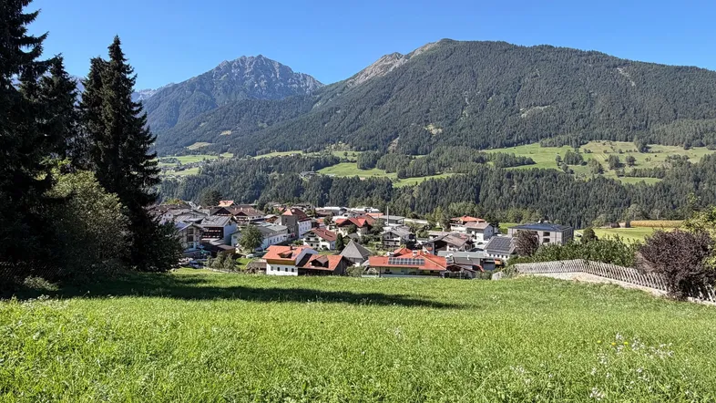 Malerisches Dorf in den Alpen, umgeben von grünen Wiesen und majestätischen Bergen unter blauem Himmel.