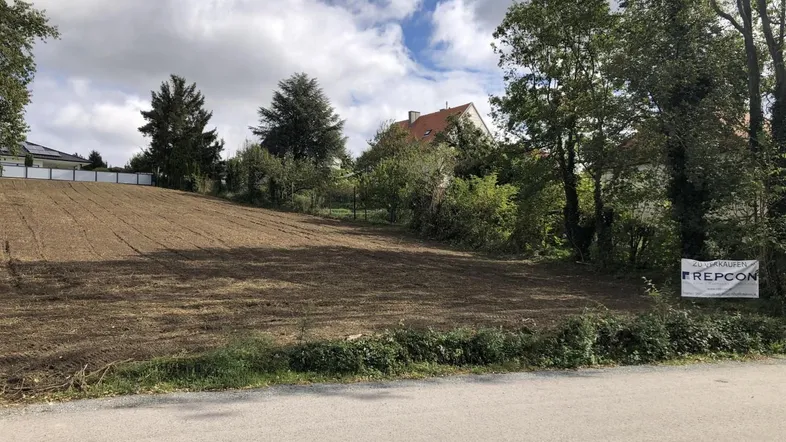 Geräumtes Grundstück mit Baustellenschild, umgeben von Bäumen und Häusern in der Ferne.