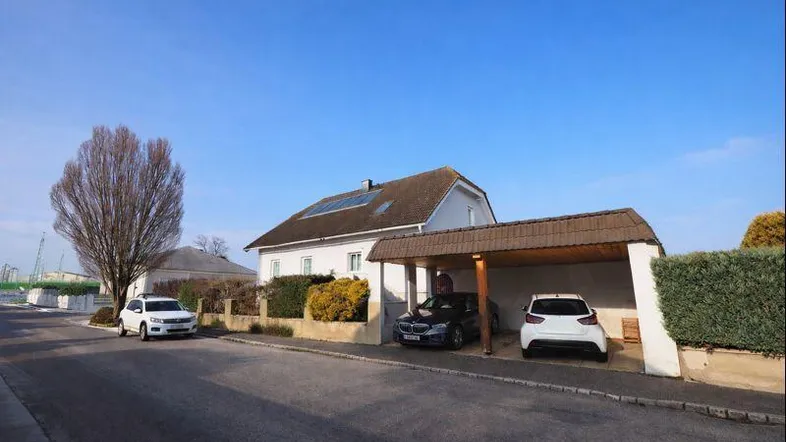 Weißes Wohnhaus mit Satteldach, Carport und Vorgarten an einer ruhigen Straße.