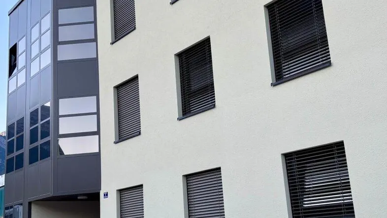 Moderne Gebäudefassade mit hellen Wänden, dunklen Fensterläden und einem gläsernen Anbau.