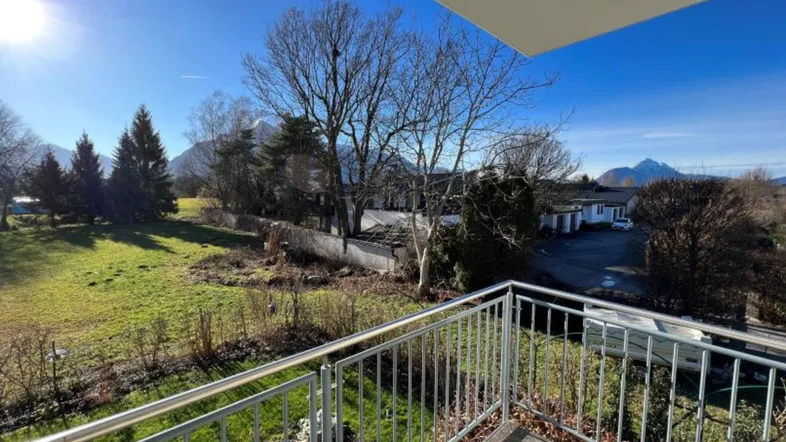 Heller Balkon mit Metallgeländer und weitem Blick auf grüne Wiesen und Berge unter blauem Himmel.