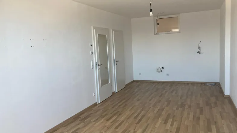 Leerer Raum mit Parkettboden, weißen Wänden und Anschlüssen für eine zukünftige Küche oder Waschküche.