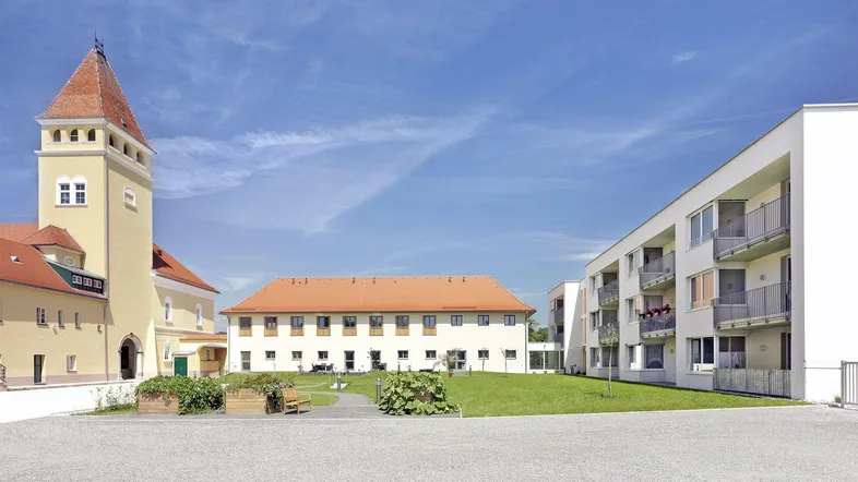 Gesamtanlage mit historischem Gebäude, modernen Wohnbauten und begrüntem Innenhof unter blauem Himmel.