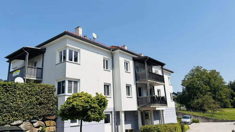 Weißes Mehrfamilienhaus mit Balkonen und gepflegter Hecke unter blauem Himmel.