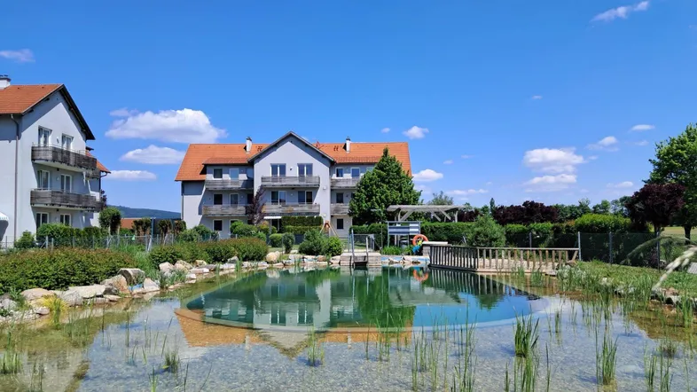 Gebäude mit mehreren Balkonen und einem natürlichen Schwimmteich in einer grünen Umgebung unter blauem Himmel.