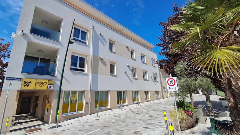 Modernes Mehrfamilienhaus mit Geschäften im Erdgeschoss und Balkonen an einer gepflasterten Straße.