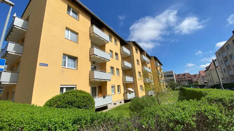 Gelbes Mehrfamilienhaus mit mehreren Balkonen und gepflegter Grünanlage unter blauem Himmel.