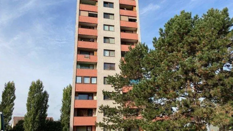 Hohes Mehrfamilienhaus mit zahlreichen Balkonen und umgebender Grünfläche unter blauem Himmel.