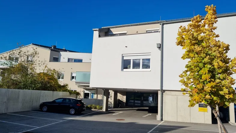 Moderne Gebäudefassade mit Außenparkplätzen und Herbstbaum unter blauem Himmel.